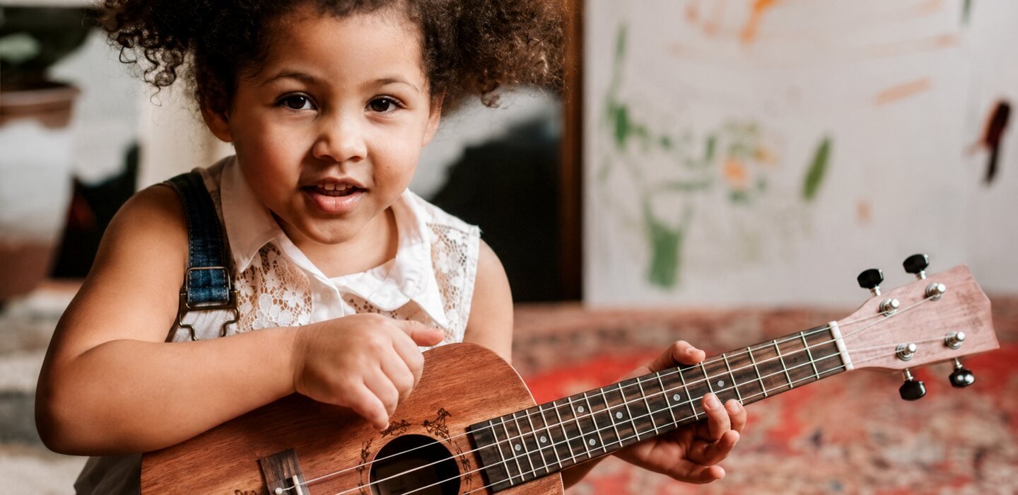 A young child sitting on a patterned rug, playing a small wooden ukulele.